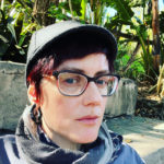 Headshot of an androgynous person with glasses, pale skin, and dark hair. They are sitting in a park with a neutral facial expression.